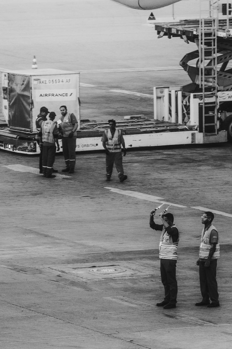 Black and white photo of airport ground crew assisting an aircraft on the tarmac with clear communication and safety measures.